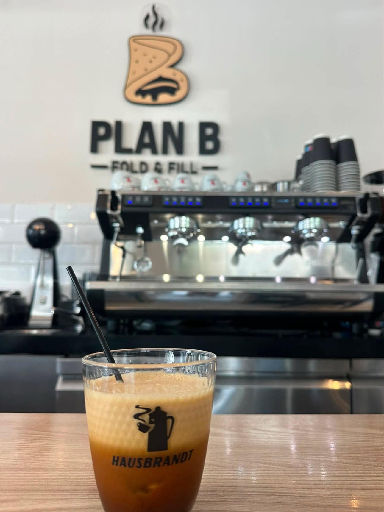 Καφές στο Plan B μπροστά από τη μηχανή espresso και το λογότυπο του καταστήματος